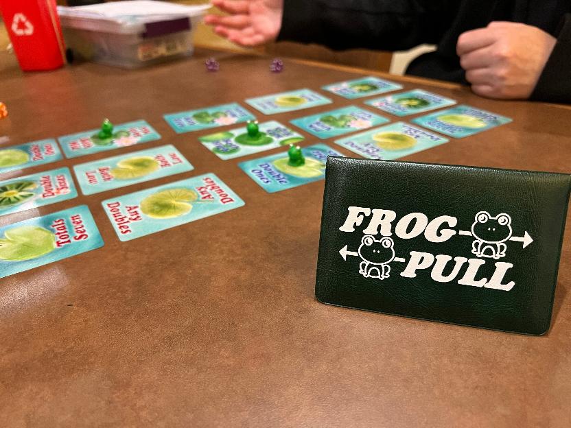 cards laid out in a grid with three resin frogs on top of them and the game wallet standing in the foreground