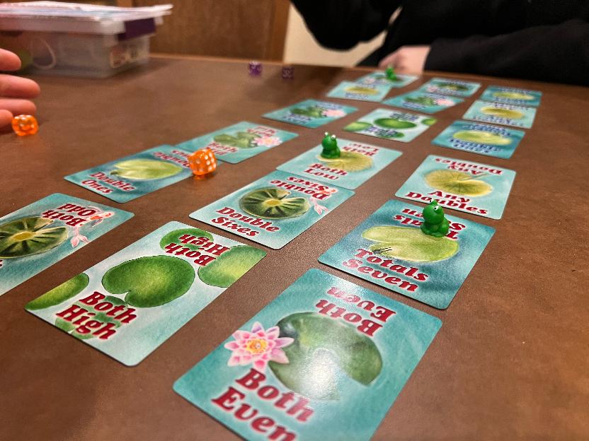 a close up angle from one end of of the cards laid in a grid. Resin frogs are on top of the cards and orange dice are mid-roll.