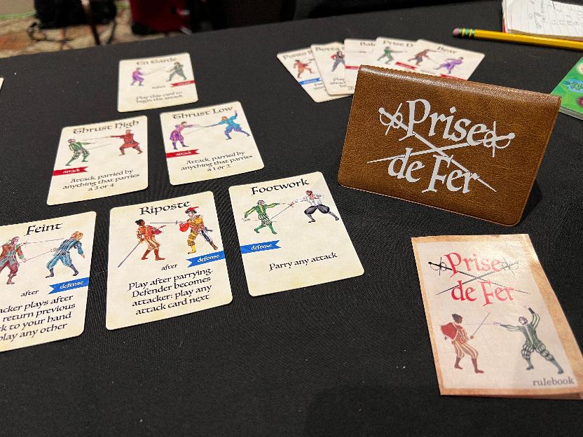 Game cards laid out in a pyramid pattern, a small fan of cards in the background, the wallet standing to the side, and instruction booklet in the foreground.